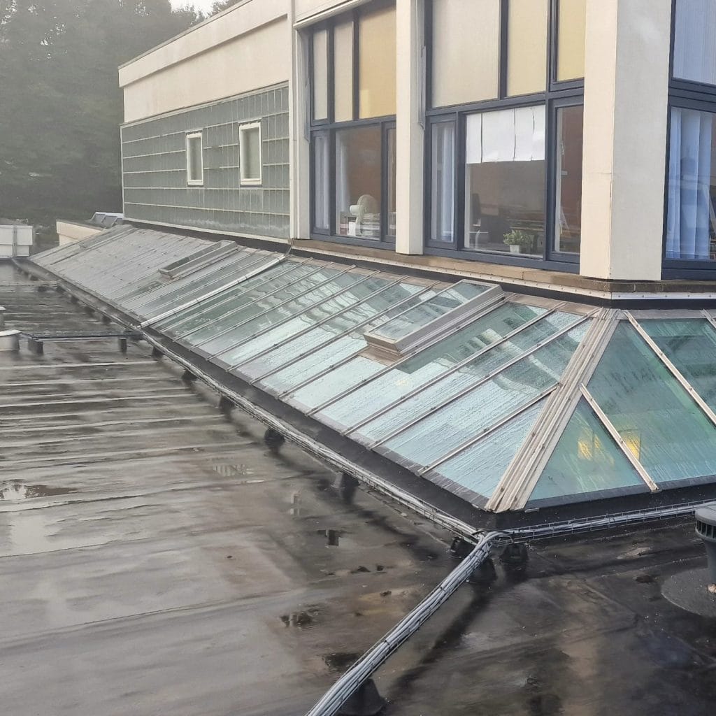 Glasreparatie en onderhoud op het platte dak van een modern gebouw met veel glasramen en dagscheiding.